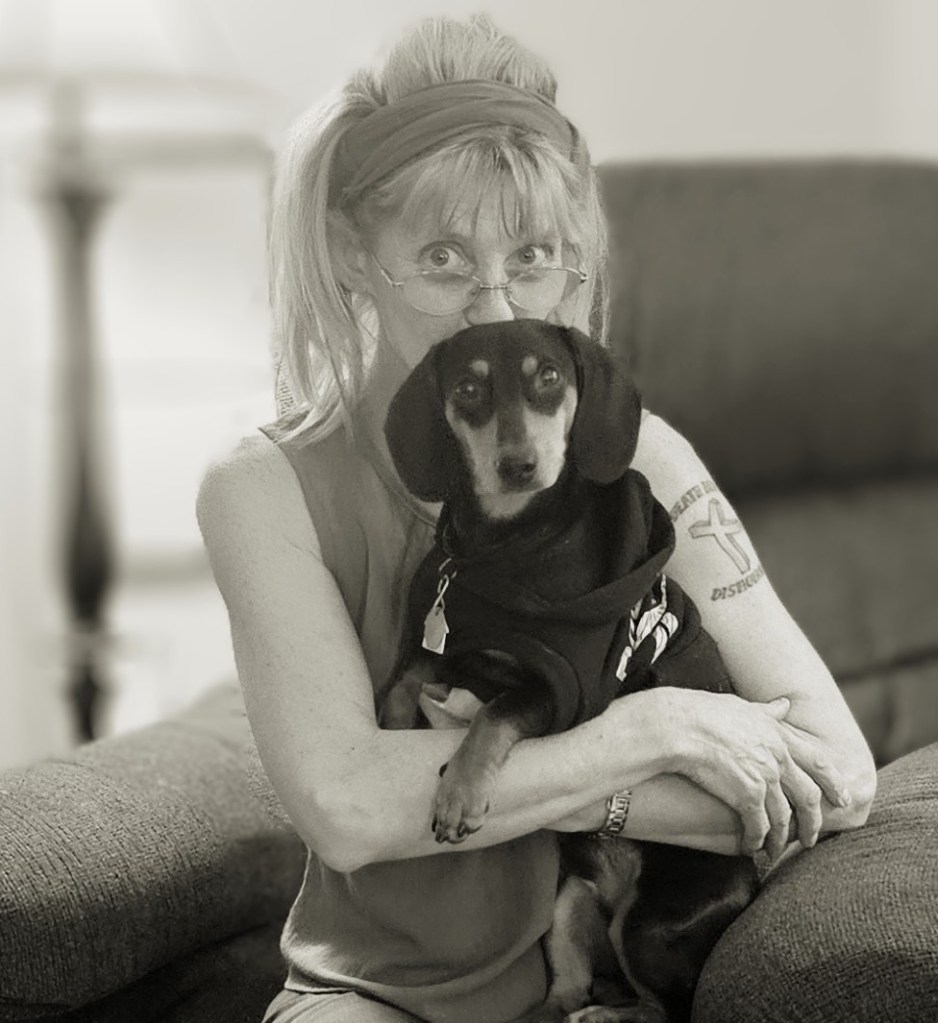 A woman with glasses and a headband sits on a couch, holding a small black and tan dog in her arms. The photo is in black and white, capturing a cozy moment.
